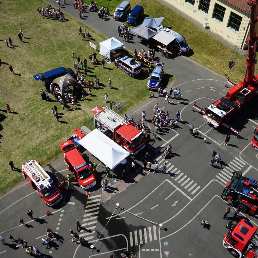 Luftaufnahme einer Versammlung mit mehreren Feuerwehrfahrzeugen, Zelten und Menschen auf einem Grasfeld und einer Straße. Mehrere Fahrzeuge sind geparkt.