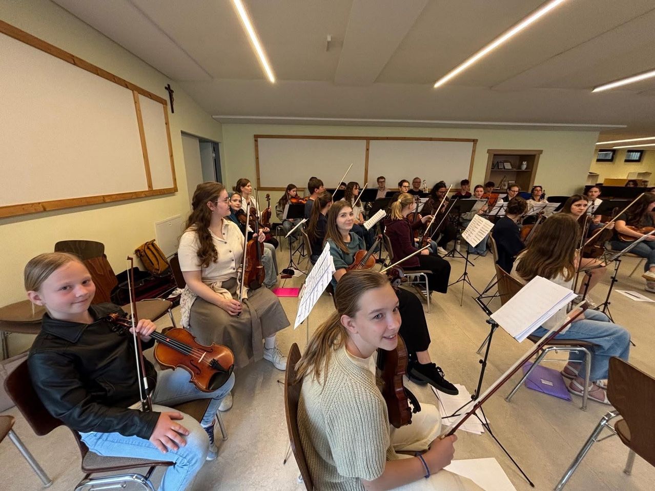 Eine Gruppe junger Musiker übt zusammen, jeder hält eine Geige. Sie sitzen in Stühlen im Raum, lesen Noten und lächeln.