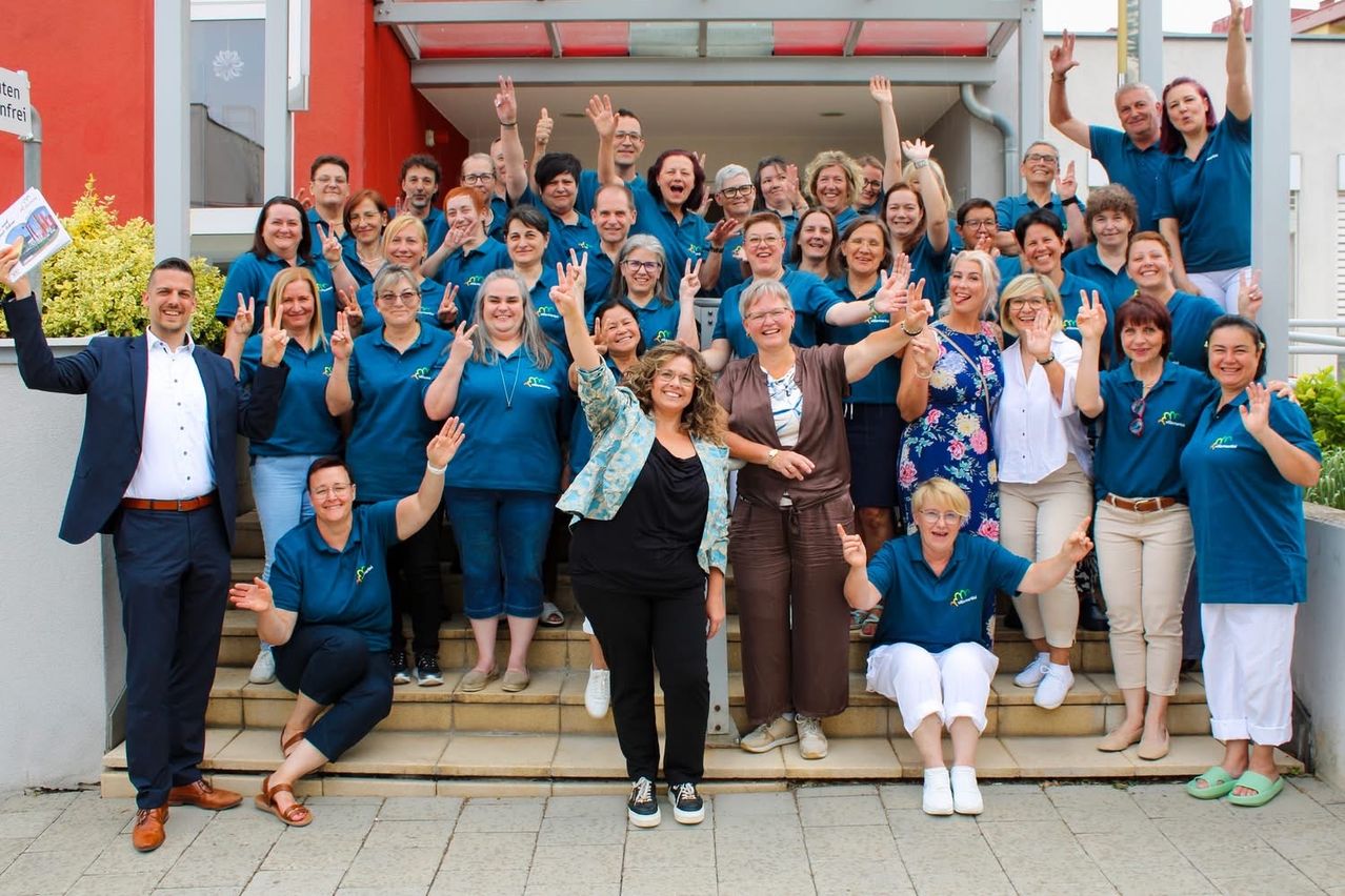 Eine Gruppe von Menschen in blauen Hemden posiert für ein Foto auf den Stufen vor einem Gebäude. Sie lächeln und machen verschiedene Handgesten.