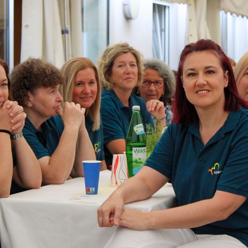 Eine Gruppe von Frauen sitzt an einem Tisch, lächelt und hört zu. Eine Flasche Walda und ein blauer Becher stehen auf dem Tisch. Die Frau rechts trägt ein blaues Shirt mit einem Logo.