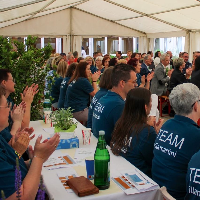 Eine Gruppe von Menschen in passenden blauen Shirts sitzt unter einem Zelt, klatscht und lächelt. Ein Tisch hat eine grüne Flasche, blaue Becher und Papiere. Ein Topf mit Pflanze steht auf dem Tisch.