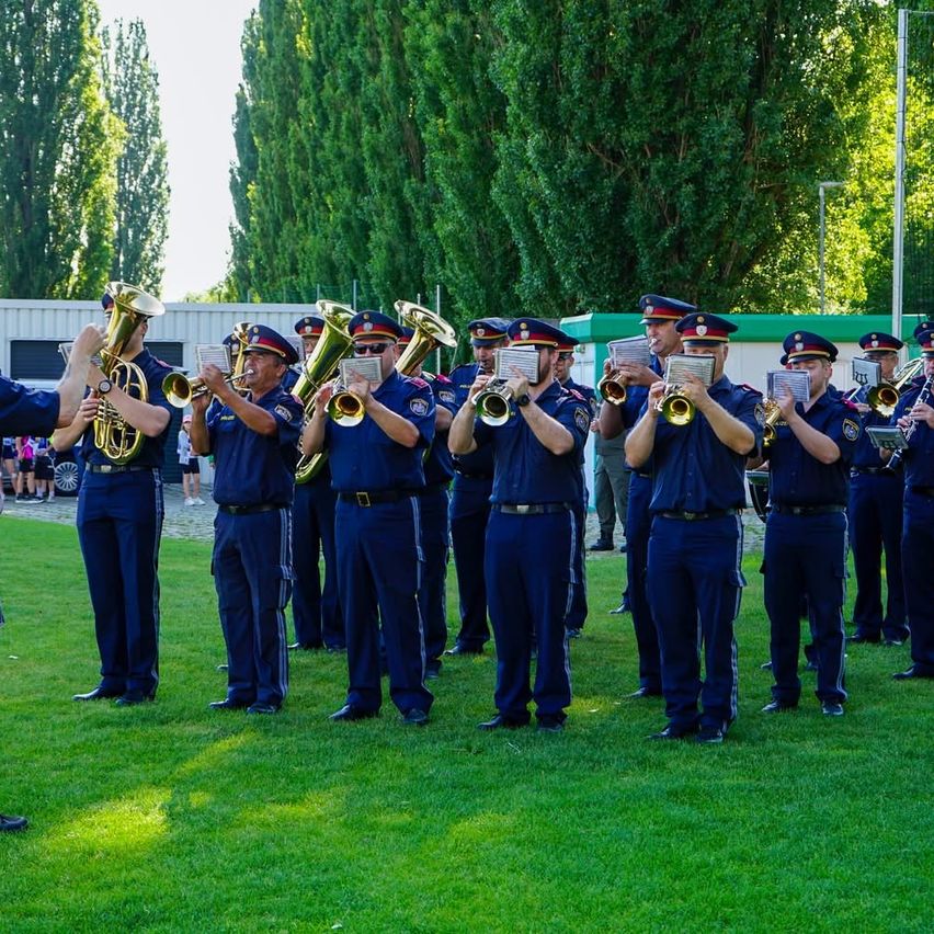 Eine Militärkapelle spielt auf einem Rasenfeld. Sie spielen Blechblasinstrumente, darunter Trompeten und Tuben. Sie stehen in Formation, einige von ihnen tragen Sonnenbrillen. Dahinter steht ein Gebäude mit grünem Rand und Bäumen.