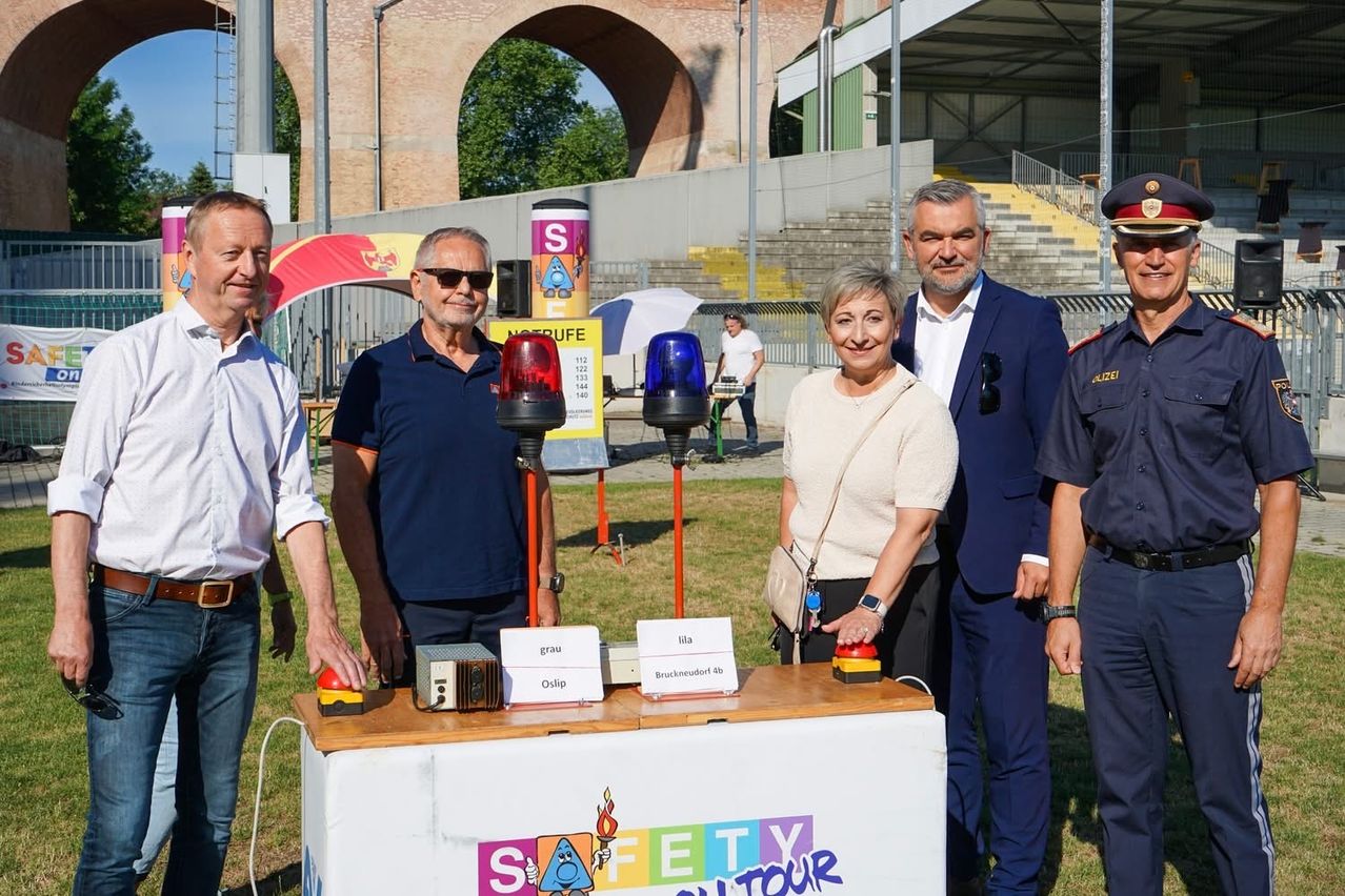 Fünf Erwachsene stehen neben einem Stand mit blinkenden Lichtern, einem Schild mit dem Wort Sicherheit und dem Wort Tour. Sie lächeln für die Kamera. Im Hintergrund sind ein Stadion und einige Personen zu sehen.