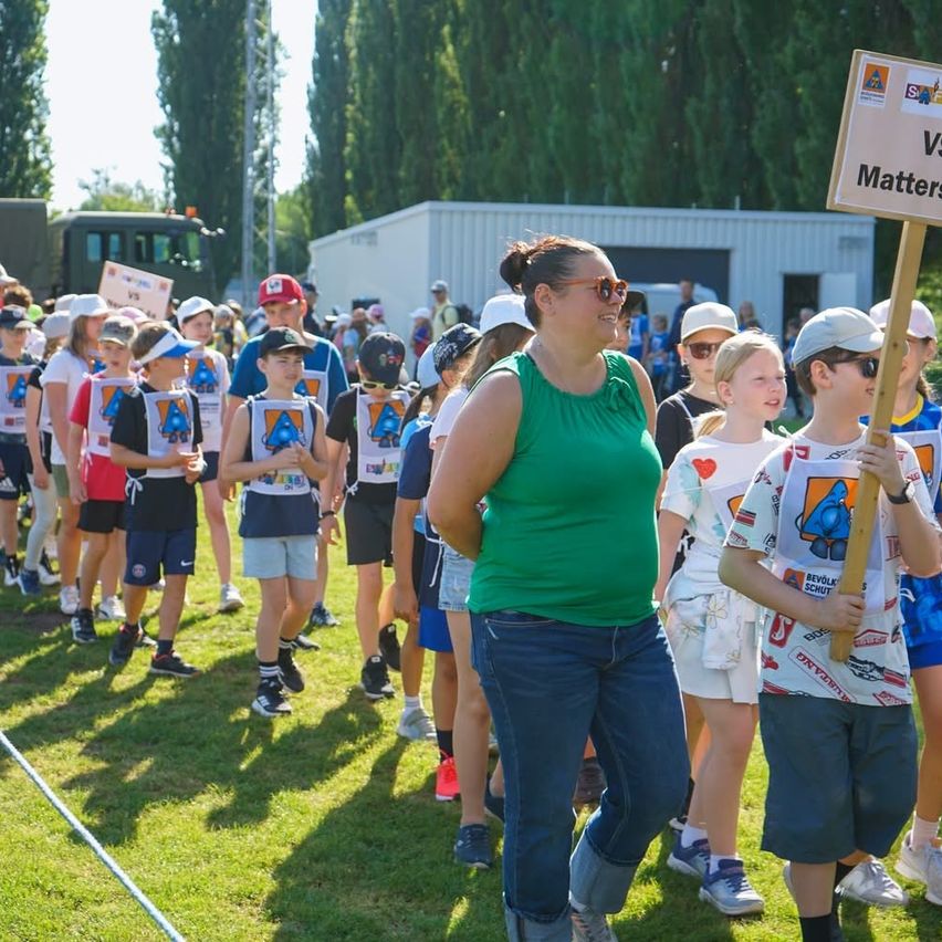 Eine Gruppe von Menschen steht auf einem Grasfeld, trägt T-Shirts mit einem Logo und einige halten Schilder. Eine Frau in Grün steht neben einem Schild, auf dem Vs Matters steht. Dahinter sind ein Gebäude und ein LKW zu sehen.