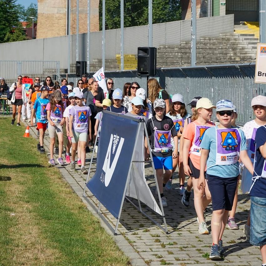Eine Gruppe von Kindern läuft auf einem gepflasterten Weg und trägt Mützen und T-Shirts. Sie stehen in einer Reihe, und einige tragen Banner. Lautsprecher sind an einem Zaun mit einem Banner aufgestellt. Bäume und ein Gebäude befinden sich hinter dem Zaun.