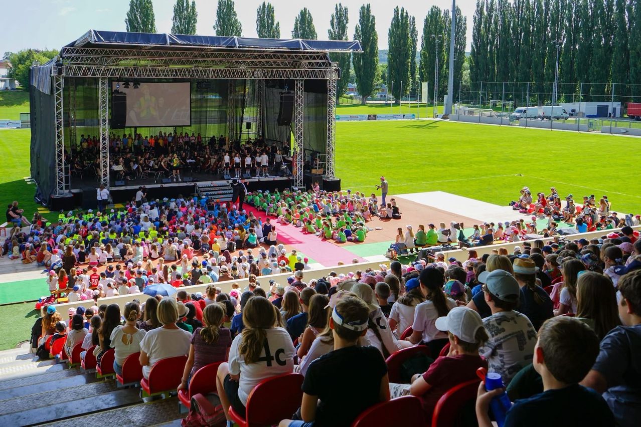 Eine große Menschenmenge versammelt sich in einem Stadion und sieht ein Orchester auf der Bühne. Das Publikum sitzt in bunten Stühlen, und ein grünes Feld erstreckt sich hinter der Bühne. Bäume und ein Van sind im Hintergrund zu sehen.