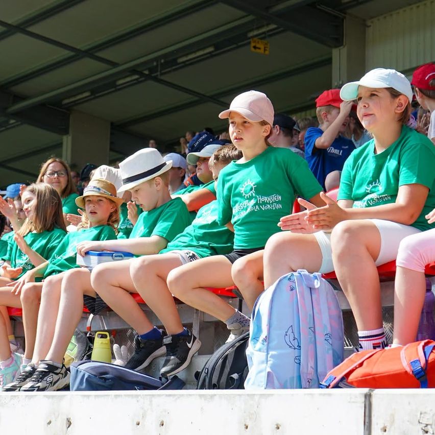 Eine Gruppe von Kindern in grünen Shirts und Mützen sitzt auf Tribünen, einige mit Brillen, und eine mit einem Rucksack. Sie beobachten wahrscheinlich eine Veranstaltung, mit anderen im Hintergrund ebenfalls sitzend.