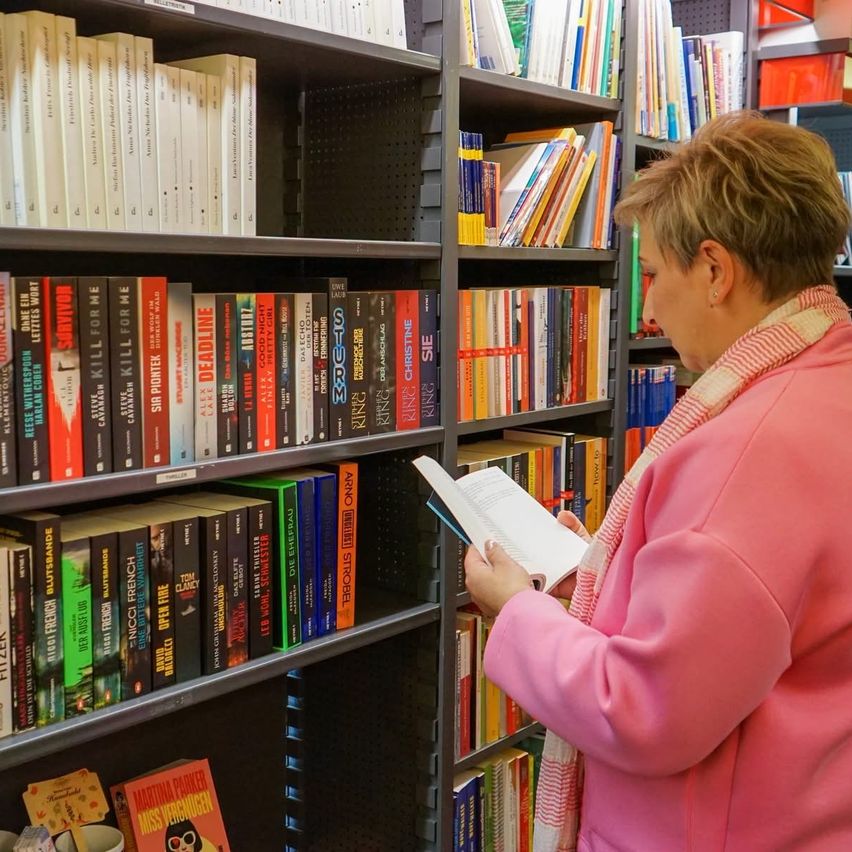 Eine Frau in einem rosa Mantel und einer Schal blättert in einem Bücherregal. Die Regale sind gefüllt mit verschiedenen Büchern, einige sind prominent mit ihren Rücken sichtbar platziert.