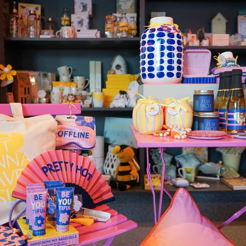 Ein farbenfrohes Ladendisplay mit verschiedenen Produkten auf Regalen und Tischen. Die Regale enthalten Flaschen, Gläser und Schachteln. Ein Tisch zeigt einen rosa Fächer, blau-weiße Gegenstände und eine Tasche. Es gibt eine Vielzahl von Artikeln, einschließlich einer Spielzeugbiene.