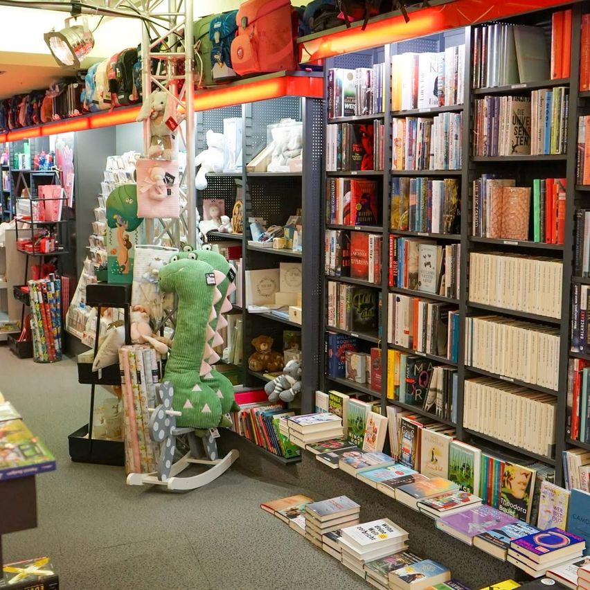 Ein Geschäft gefüllt mit Büchern, Spielzeug und Kuscheltieren. Regale mit Büchern, Plüschtieren und einem Schaukelstuhl. Taschen und Artikel sind auf Regalen ausgestellt.