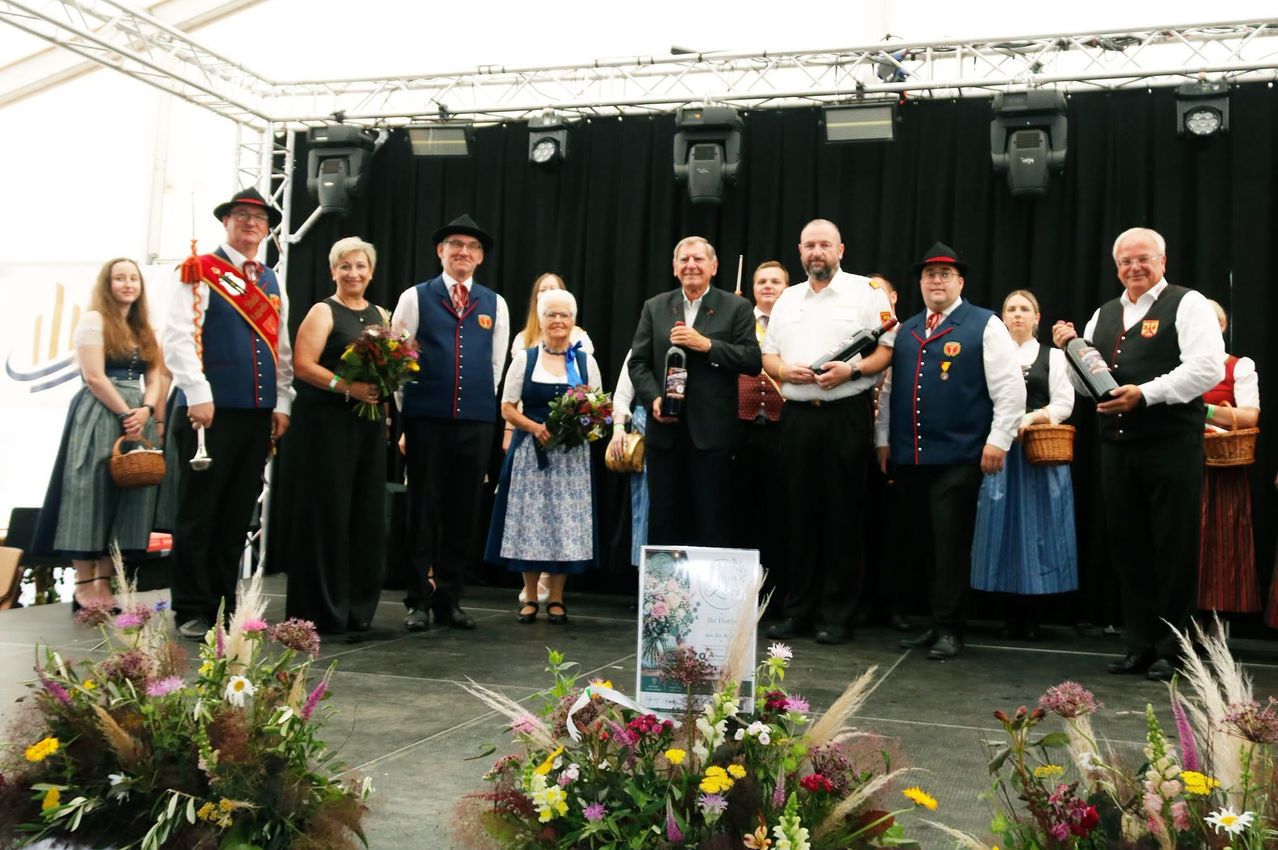 Eine Gruppe von Menschen steht auf der Bühne, hält Blumen und Weinflaschen. Sie tragen traditionelle Kleidung, und ein Schild ist auf dem Boden. Scheinwerfer sind über ihnen.
