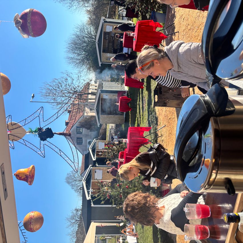 Eine Gruppe von Menschen ist unter blauem Himmel im Freien versammelt. Eine Frau schenkt Getränke aus einem Krug ein. Zwei Personen lehnen sich an einem Griller an. Ein roter Ballon schwebt oben. Es gibt einige kleine Häuser und einen Baum.