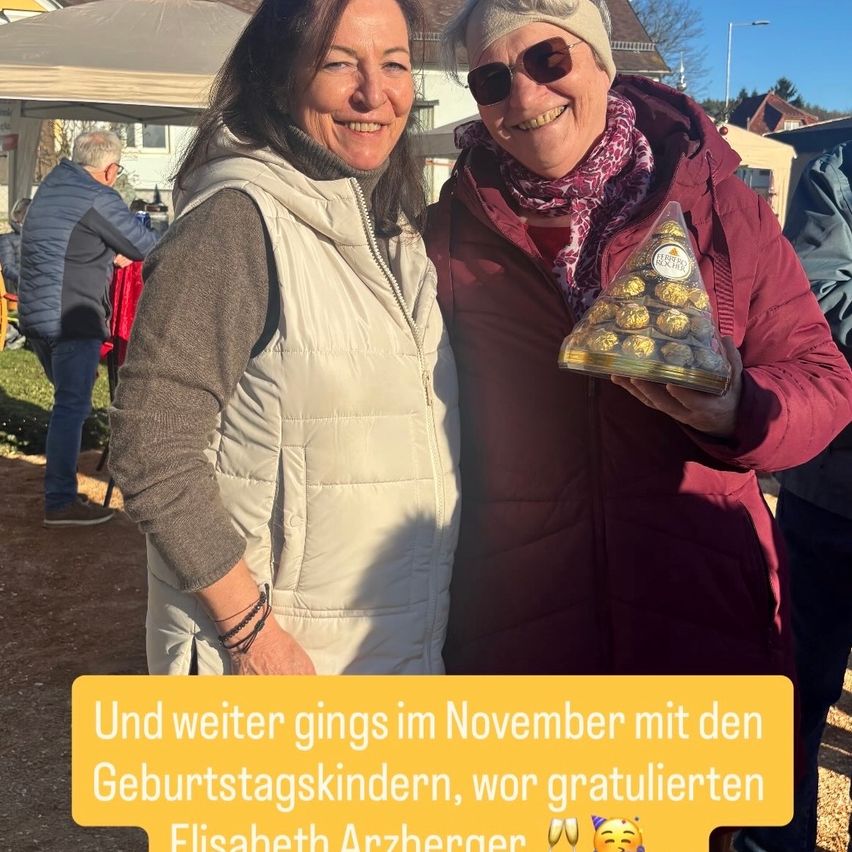 Zwei lächelnde Frauen, eine hält ein dreieckiges Paket mit Schokoladen, stehen draußen. Ein Text-Overlay lautet: 'Und weiter ging es im November mit den Geburtstagskindern, die Elisabeth Arzhberger gratulierten.'