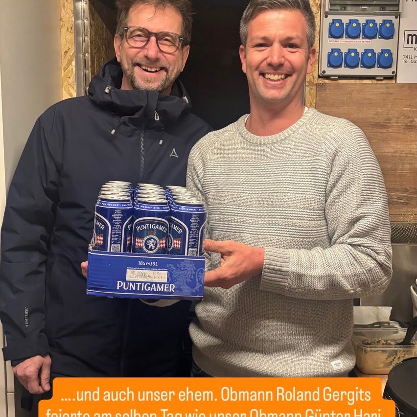 Zwei lächelnde Männer, einer hält eine Bierkiste in der Hand, mit einem Untertitel auf Deutsch, der besagt: '...und auch unser ehemaliger Chef Roland Gergits feierte am selben Tag wie unser Chef Gunter Hari.'