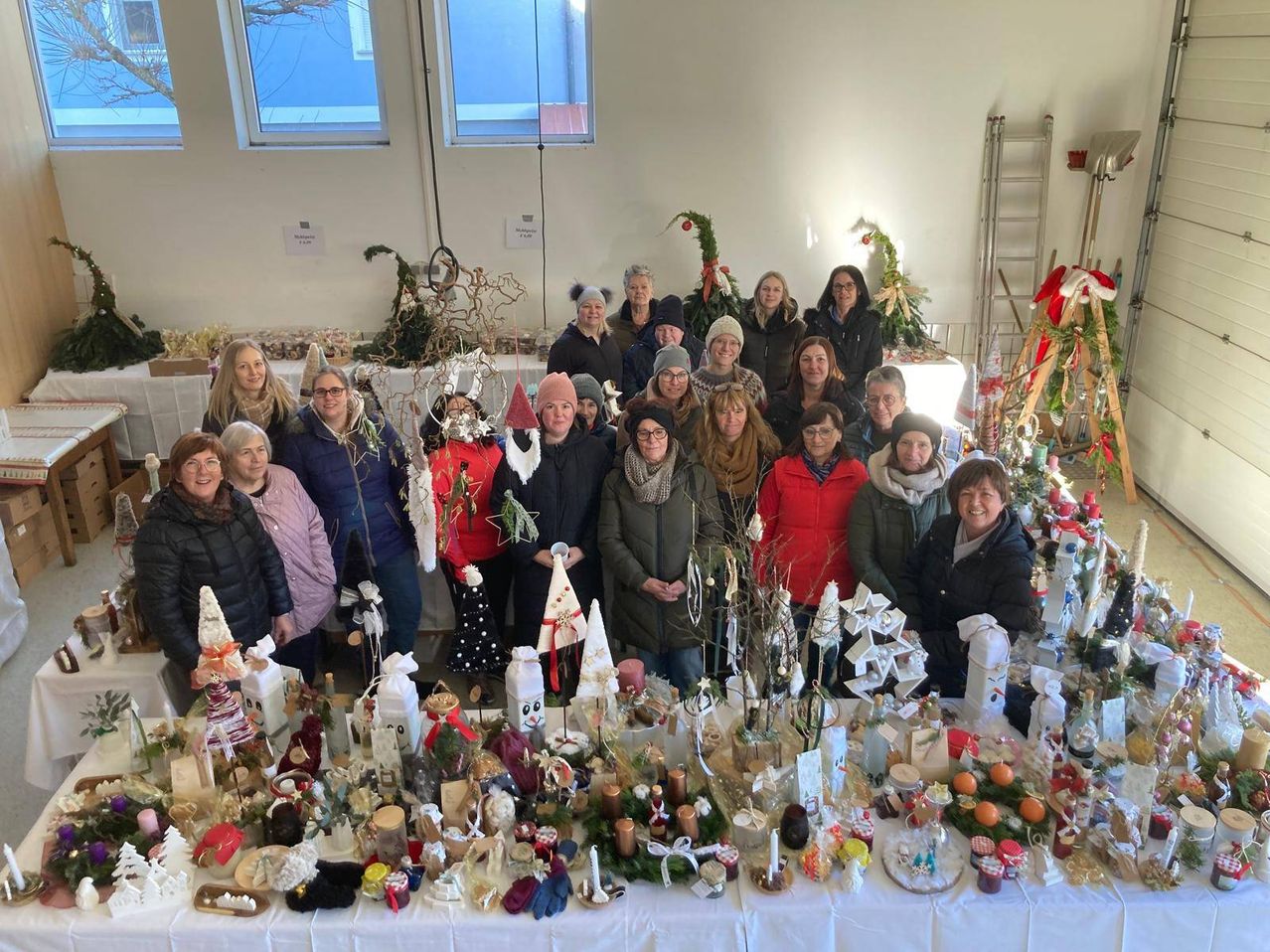Eine Gruppe von Frauen steht hinter einem Tisch voller Weihnachtsdekoration. Sie lächeln und posieren für ein Foto. Es gibt Weihnachtsbäume, Ornamente und andere festliche Gegenstände auf dem Tisch.