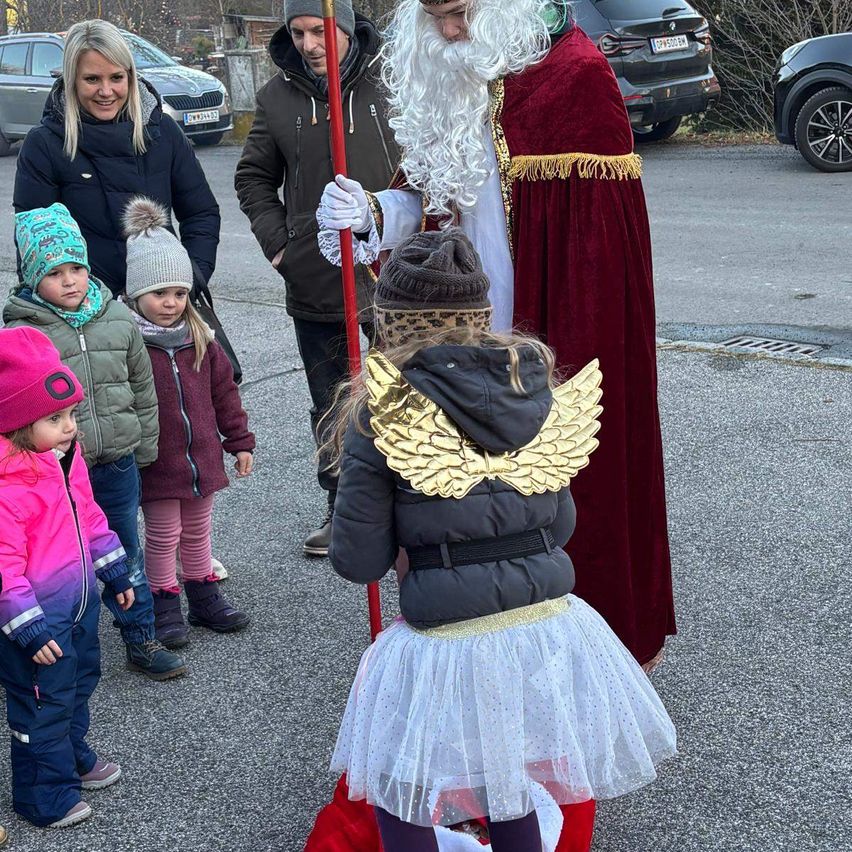 Eine Gruppe von Kindern und Erwachsenen steht auf einem Parkplatz. Ein Mann in Santa Claus-Kostüm hält einen Stab. Ein Mädchen in einem Engelkostüm steht vor ihm. Dahinter sind Autos geparkt.
