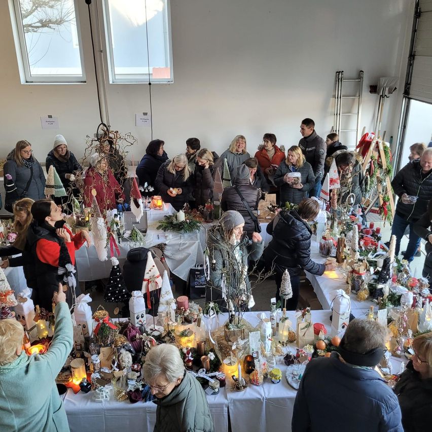 Ein Raum voller Menschen rund um Tische mit Kerzen, Ornamenten und anderen Gegenständen. Sie diskutieren möglicherweise etwas, während sie die Gegenstände auf den Tischen betrachten. Einige Menschen tragen Hüte und Brillen. Der Raum hat weiße Wände und Glasfenster. In der Nähe der Fenster befinden sich Leitern.