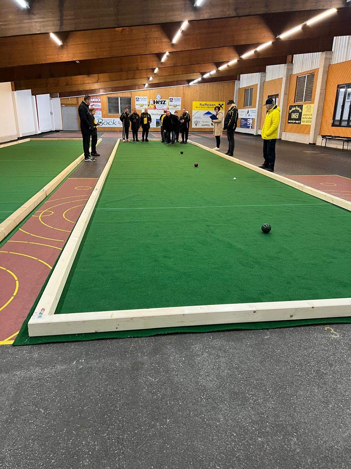 Eine Gruppe von Menschen steht um einen grünen Rasen herum, der Teil eines großen Innenfeldes ist, möglicherweise für ein Spiel wie Curling. Einige Menschen stehen in einer Reihe in der Nähe der Wand.