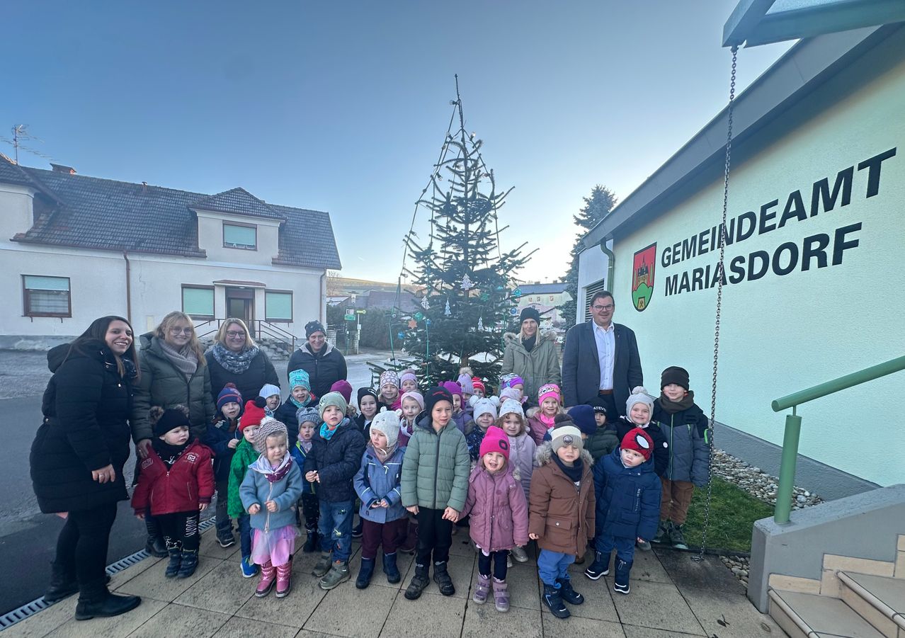 Eine Gruppe von Kindern und Erwachsenen steht vor einem Weihnachtsbaum. Dahinter befindet sich ein Gebäude mit der Aufschrift Gemeinde Mariasdorf an der Wand.