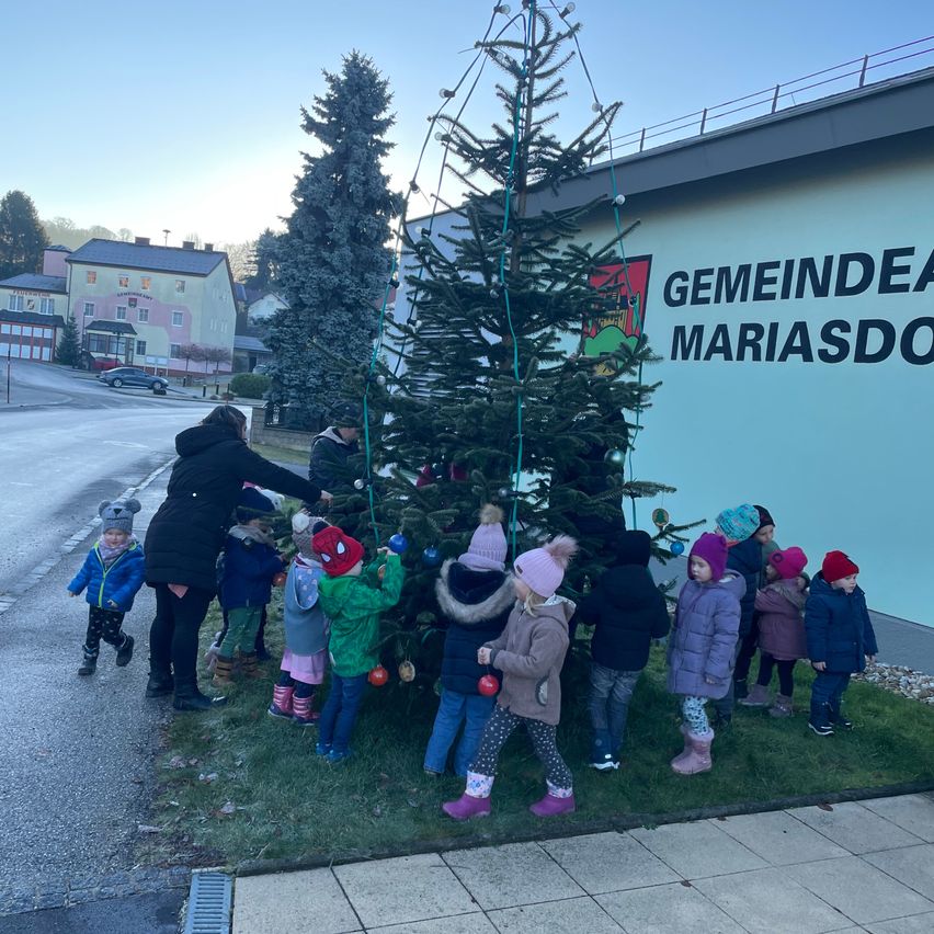 Eine Gruppe von Kindern und einem Erwachsenen schmückt eine Weihnachtsbaum vor einem Gebäude mit dem Schild Gemeinde Mariasdorf.