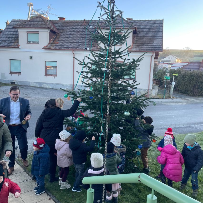 Eine Gruppe von Kindern und Erwachsenen schmückt einen hohen Weihnachtsbaum vor einem Haus an einem sonnigen Tag.