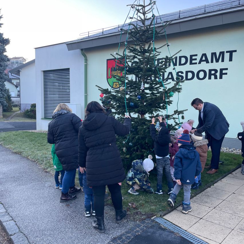 Eine Gruppe von Kindern und Erwachsenen schmückt einen Weihnachtsbaum vor einem Gebäude namens Endamt Hasdorf.