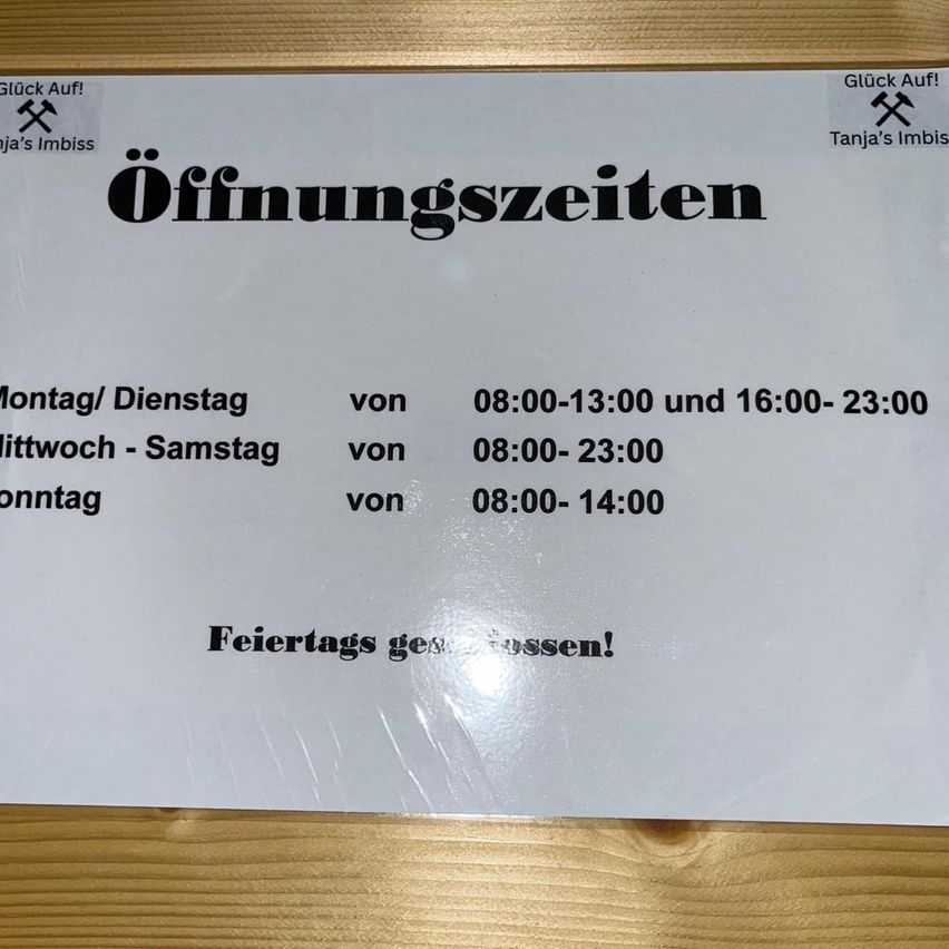 Ein weißes Schild zeigt die Öffnungszeiten von Tanjas Imbiss. Der Zeitplan zeigt, dass es von Montag bis Freitag von 8 Uhr bis 13 Uhr und von 16 Uhr bis 23 Uhr und an Wochenenden von 8 Uhr bis 14 Uhr geöffnet ist. An Feiertagen geschlossen.