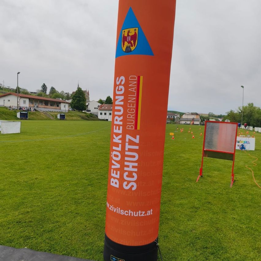 Ein hoher orangefarbener Pfahl mit den Worten 'Burgenland' und 'Bevolkerungsschutz' steht auf einem Rasenfeld mit mehreren Verkehrskegel. Hinter dem Pfahl befinden sich Häuser und ein weißes Zelt. Bäume und Straßenlaternen sind in der Ferne zu sehen.