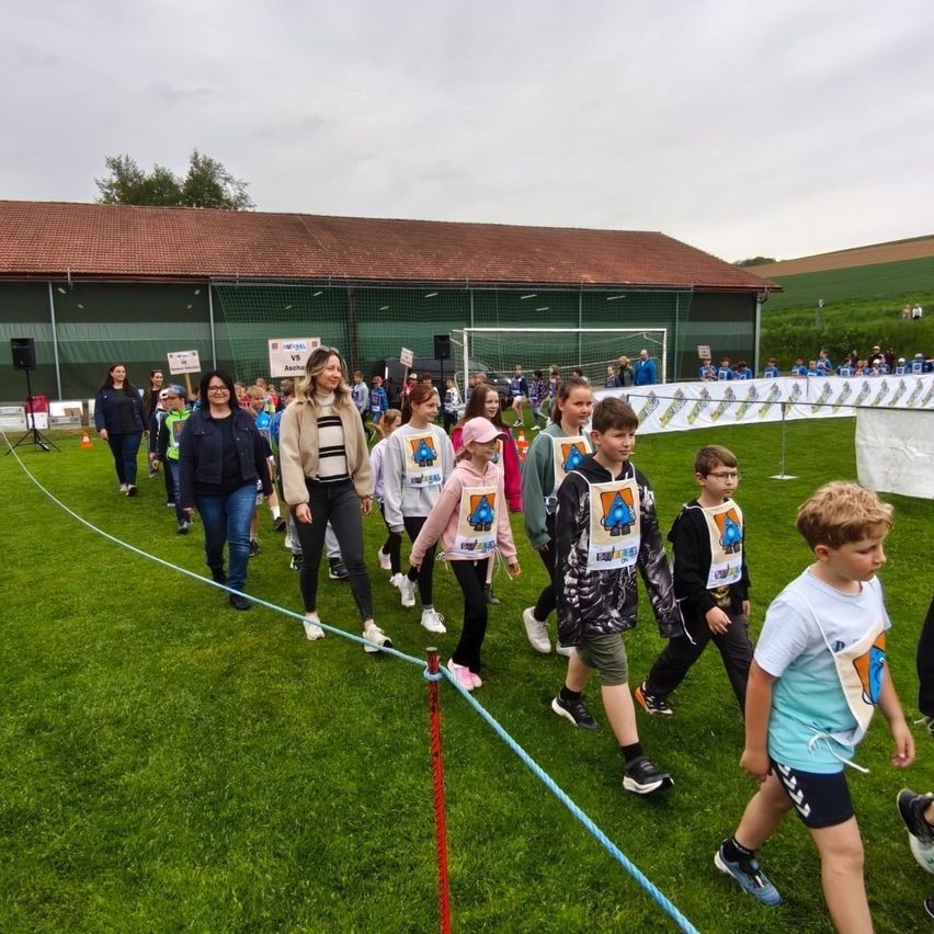 Eine Gruppe von Kindern und Erwachsenen läuft auf einem Grasfeld mit einer blauen Schnur. Sie tragen Turnschuhe und haben Rucksäcke. Die Frau vorne trägt eine Jacke und eine Brille. Dahinter steht ein Gebäude mit einem Netz und einem Fußballtor.