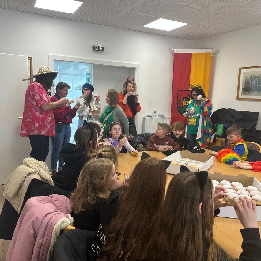 A group of people, including adults and children, are gathered in a room. Some are eating pastries from a box, while others stand or sit. A flag hangs on the wall.
