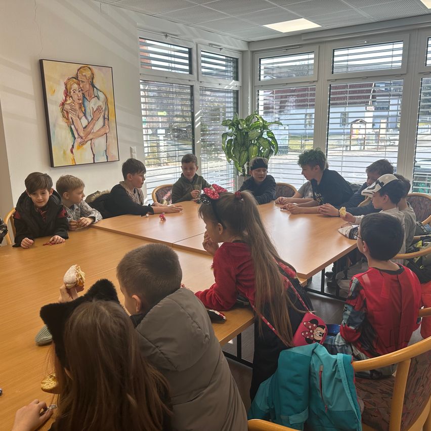 Children sit around a long table in a room with large windows, a painting on the wall, and a potted plant. Some children eat food.