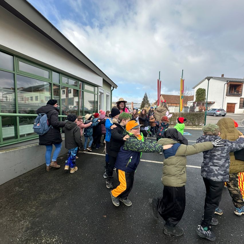 A group of children and adults in winter clothing, including hats and scarves, form a circle in an outdoor area. Some wear boots, and there is a building with glass windows in the background.