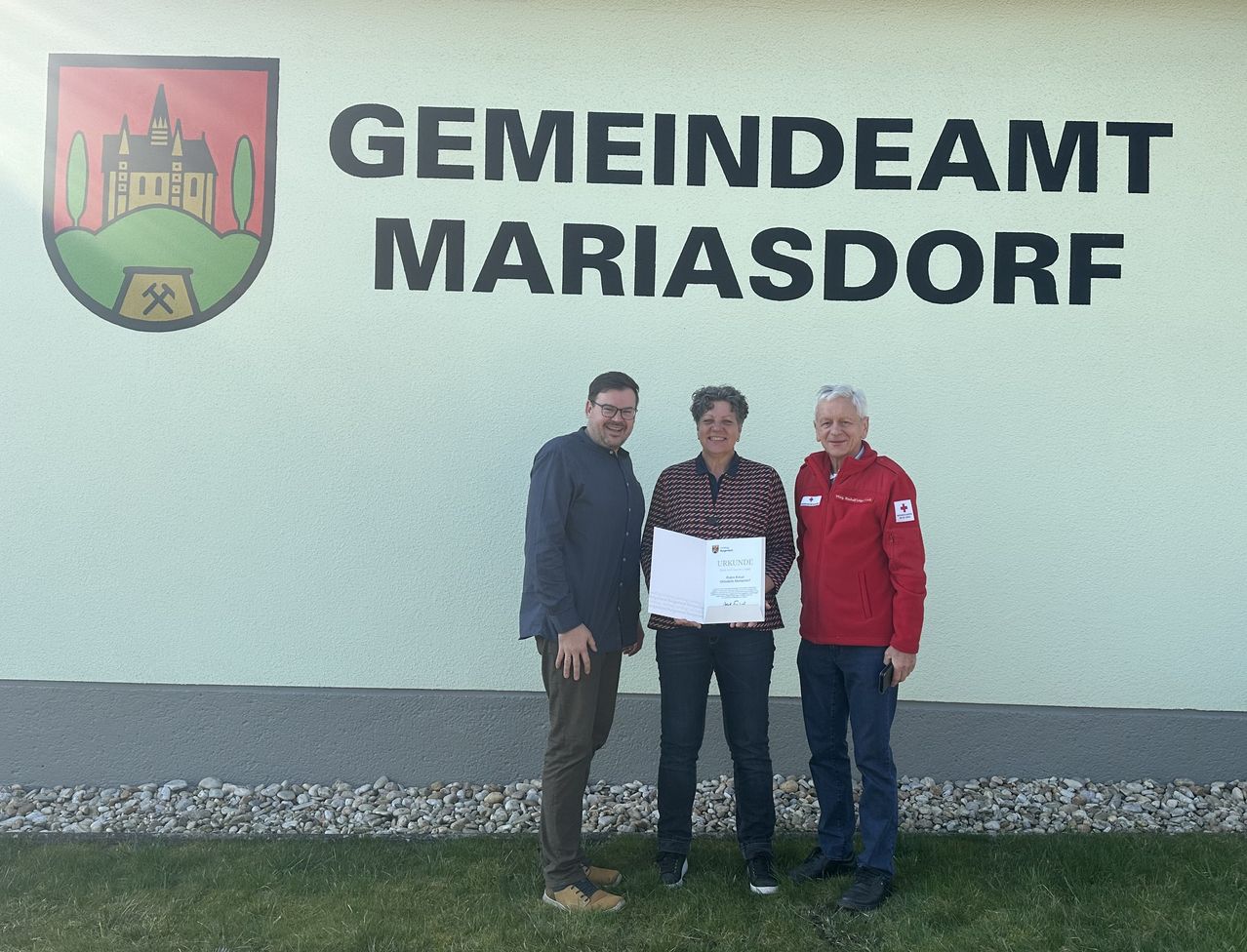 Drei Personen stehen vor einer Wand mit der Aufschrift 'Gemeindeamt Mariasdorf' und halten ein Zertifikat in der Hand. Sie lächeln.