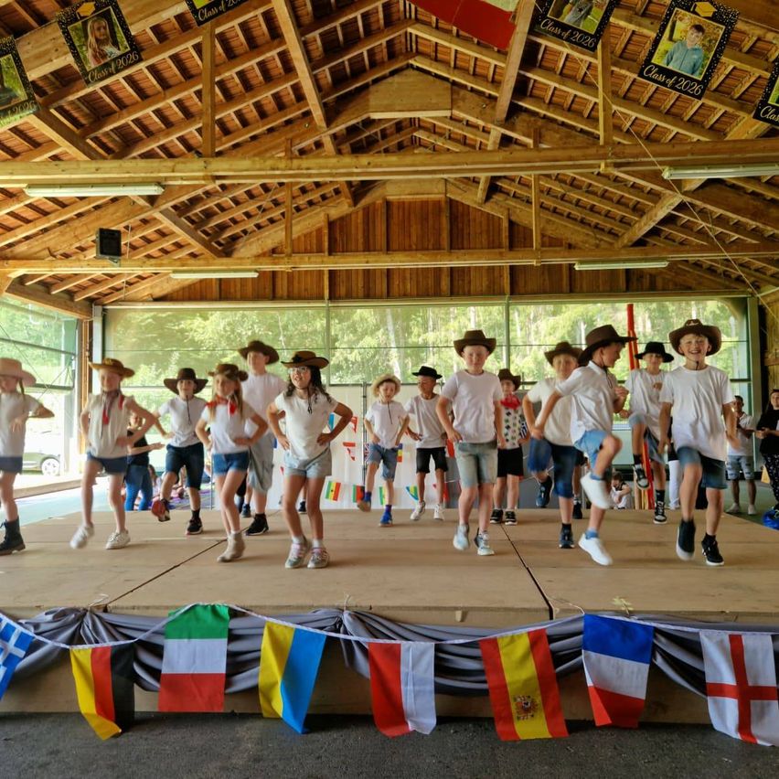 Eine Gruppe von Kindern tanzt auf einer Bühne mit Flaggen verschiedener Länder, die an der Seite hängen. Sie tragen Hüte und Turnschuhe. Im Hintergrund sitzen Leute.