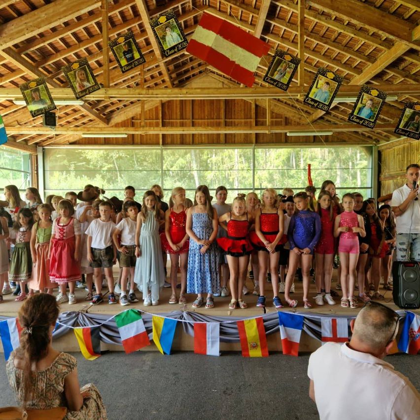 Eine Gruppe von Kindern in verschiedenen Outfits steht auf der Bühne unter einem hölzernen Dach mit Flaggen und Bannern. Ein Erwachsener steht vorne.