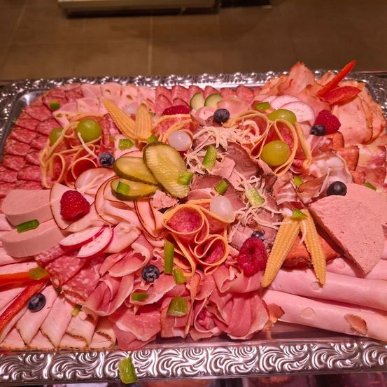 Ein silbernes Tablett enthält verschiedene Fleischsorten wie Schinken, Salami und Würste, zusammen mit eingelegtem Gemüse und Früchten wie Trauben, Beeren und Mais.