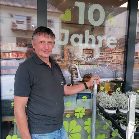 Ein Mann in einem schwarzen Shirt und Jeans steht neben einer Glastür eines Geschäfts und lächelt. Die Glastür hat ein grünes Kleeblatt-Design und den Text '10 Jahre'. Hinter ihm zeigt eine Reflexion ein Auto und ein Gebäude mit vielen Produkten.