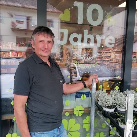 Ein Mann in einem schwarzen Shirt und Jeans steht neben einer Glastür eines Geschäfts und lächelt. Die Glastür hat ein grünes Kleeblatt-Design und den Text '10 Jahre'. Hinter ihm zeigt eine Reflexion ein Auto und ein Gebäude mit vielen Produkten.