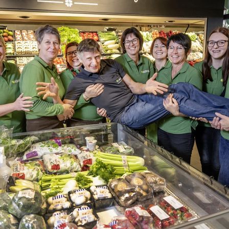 Eine Gruppe von Mitarbeitern in grünen Uniformen posiert in einem Supermarkt, einer der Männer wird von anderen hochgehoben, in der Nähe einer Ausstellung von frischem Gemüse und Obst stehend.