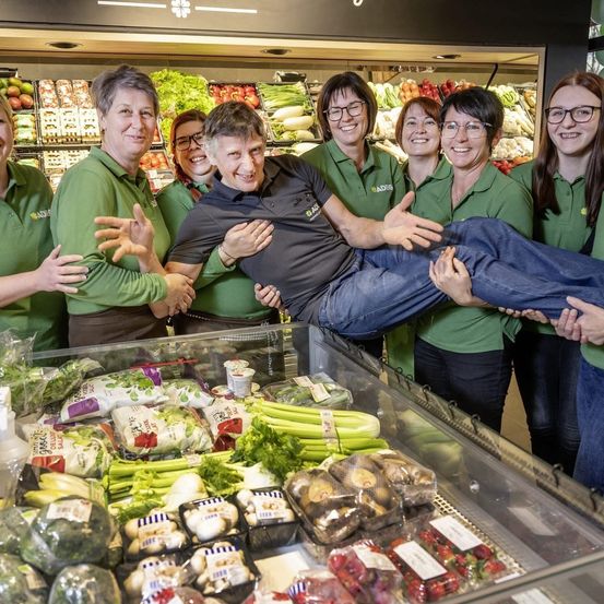 Eine Gruppe von Mitarbeitern in grünen Uniformen posiert in einem Supermarkt, einer der Männer wird von anderen hochgehoben, in der Nähe einer Ausstellung von frischem Gemüse und Obst stehend.