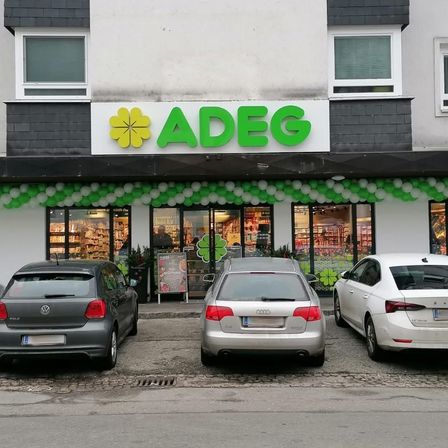 Ein Geschäft mit ADEG-Beschilderung hat grüne Ballons, die den Eingang schmücken. Drei Autos sind davor geparkt.