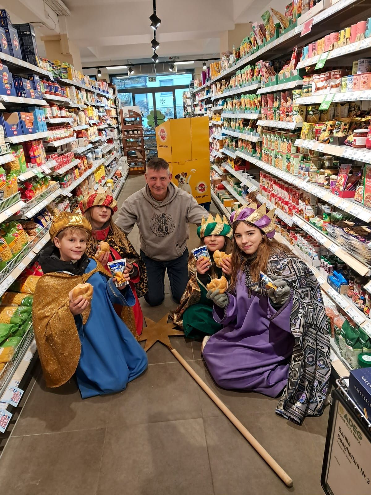 Ein Mann und vier Kinder, die als Könige gekleidet sind, posieren für ein Foto in einem Lebensmittelgeschäftsgang und halten Essen in den Händen und lächeln.