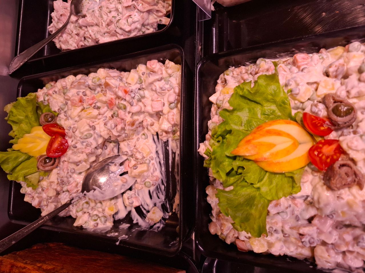 Four trays of a dish with vegetables and a creamy sauce, garnished with lettuce and sliced tomato. Each tray has a spoon on the side.