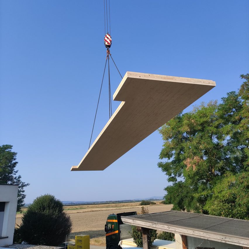 Ein großer Holzbalken wird von einem Kran unter einem klaren blauen Himmel angehoben. Bäume und ein Gebäude sind im Hintergrund.