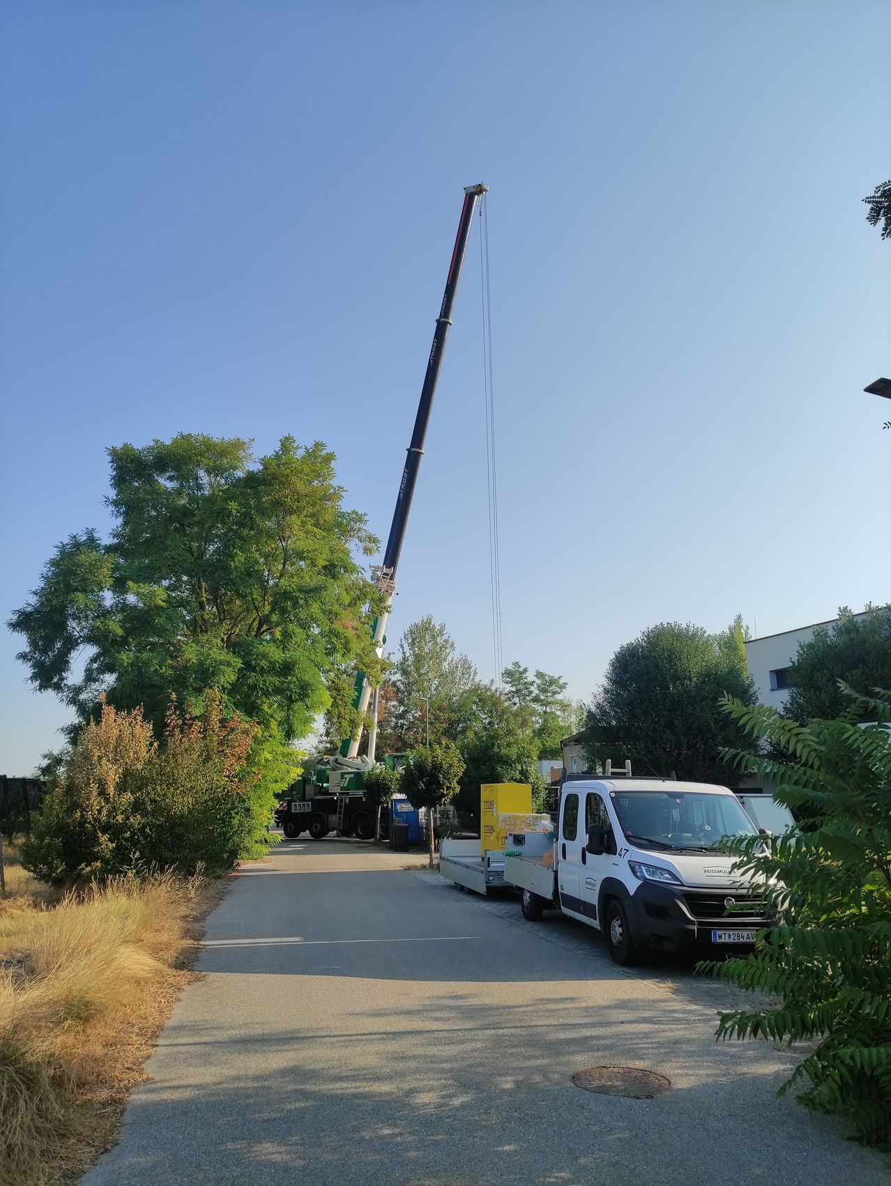 Ein Kran steht auf einer Straße, mit einem weißen Lastwagen in der Nähe. Der Kran ist nach oben ausgestreckt, und ein gelber Container ist daran befestigt. Die Szene spielt vor dem Hintergrund von Bäumen und einem Gebäude.