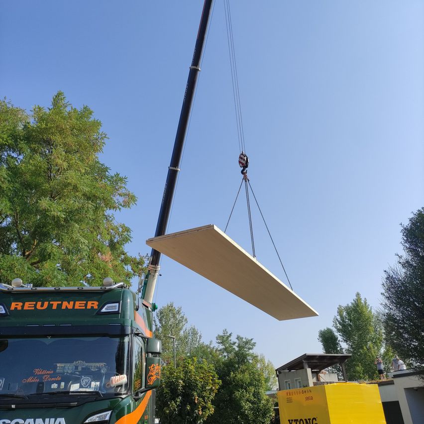 Ein großes flaches Panel wird von einem Kran von einem grünen Lastwagen mit der Aufschrift 'Reutner' angehoben. Das Panel wird über einen gelben Lastwagen mit der Aufschrift 'Ytong' bewegt. Die Szene spielt sich an einem klaren blauen Himmel mit Bäumen im Hintergrund ab.
