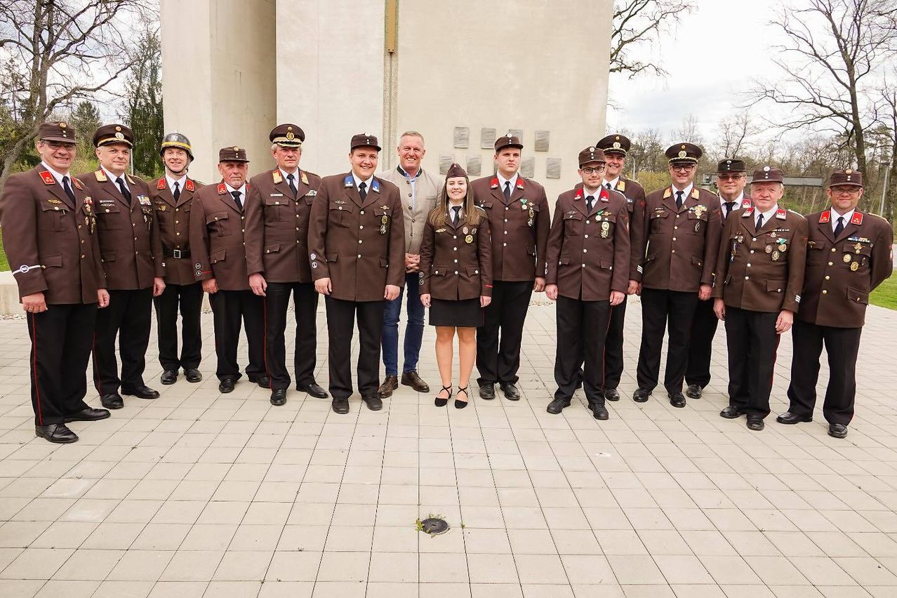 Eine Gruppe von Menschen in Militäruniformen posiert für ein Foto vor einem Denkmal, wobei eine Frau in der Mitte lächelt und von Männern umgeben ist.