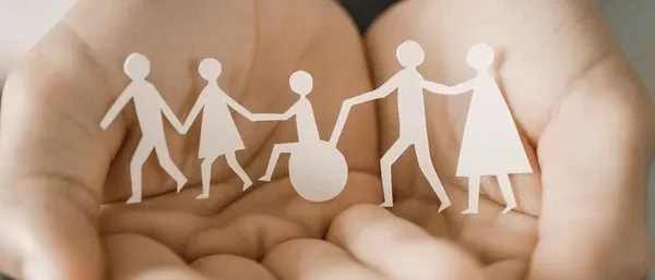 Eine Hand hält Papierausschnitte von vier Figuren, eine davon im Rollstuhl, was Inklusivität und Unterstützung symbolisiert.