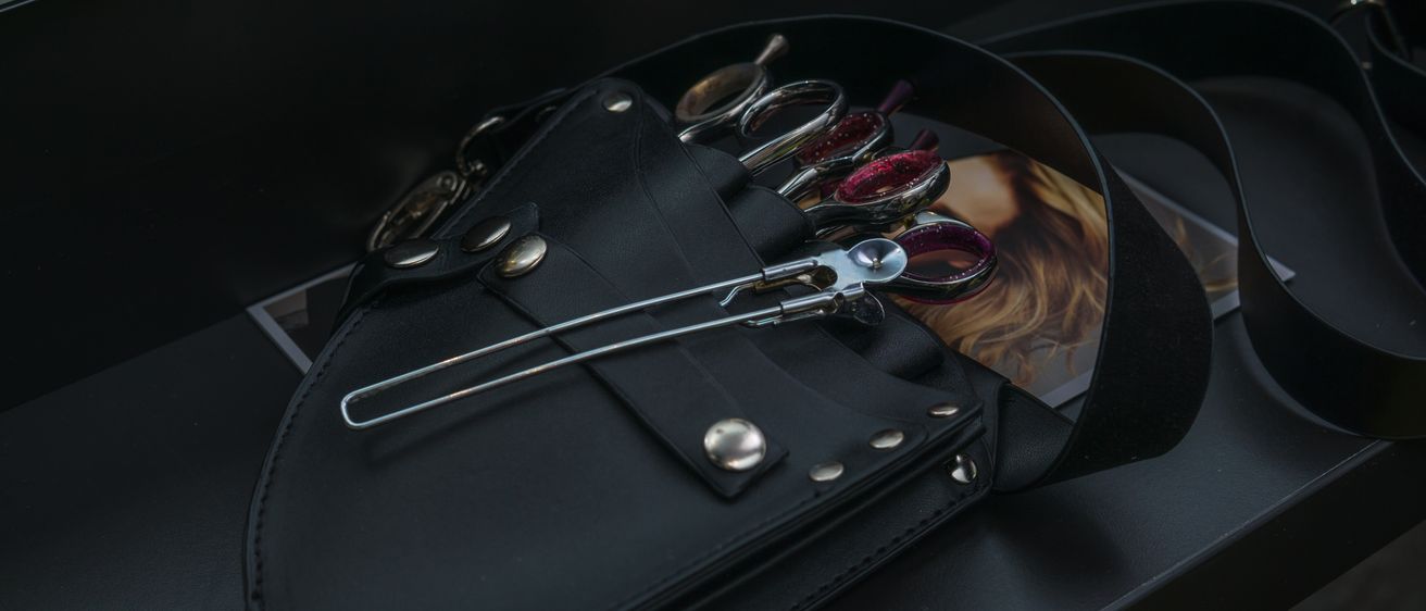 Ein schwarzer Leder-Barbierbeutel mit mehreren Scheren und einem Haarschneider im Inneren, auf einem dunklen Hintergrund präsentiert.