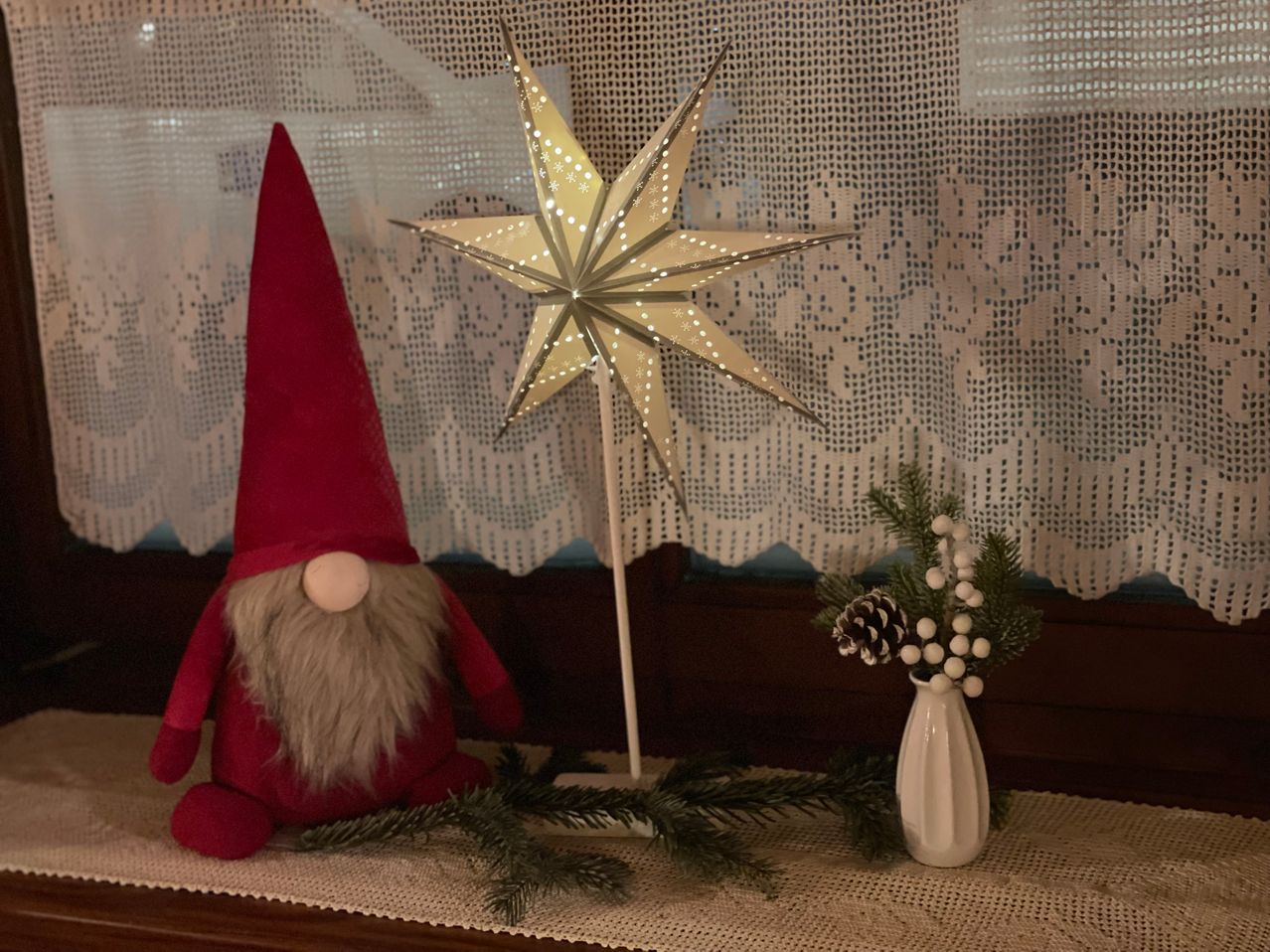 Ein roter Gnom mit weißem Bart sitzt neben einem sternförmigen Licht auf einem Tisch. Ein weißer Vase mit Tannenzapfen und Beeren steht rechts.